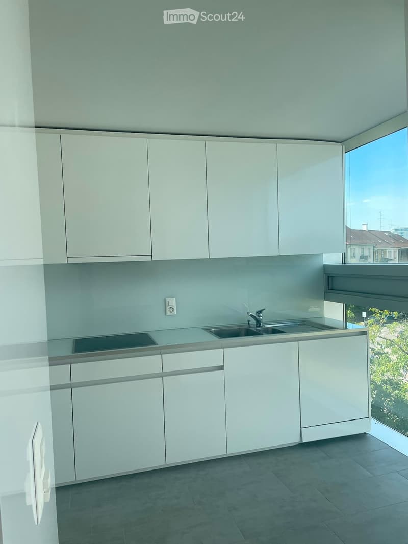 Modern kitchen with white cabinets, light grey countertops, and a double sink. Large windows provide natural light and a view of the outdoors.