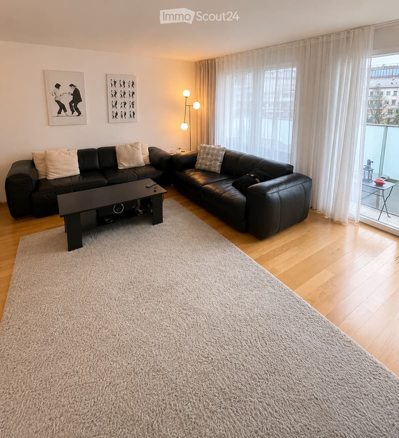 Modern living room with a black sectional sofa, coffee table, floor lamp, and large windows with sheer curtains. The room features a light-colored shag rug on a wooden floor and a small table near the balcony.