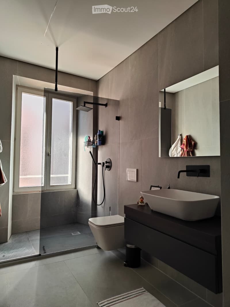 Modern bathroom with a sleek sink, toilet, glass-enclosed shower, and large mirror. Features include a dark countertop, modern faucet, and built-in shelving with toiletries.