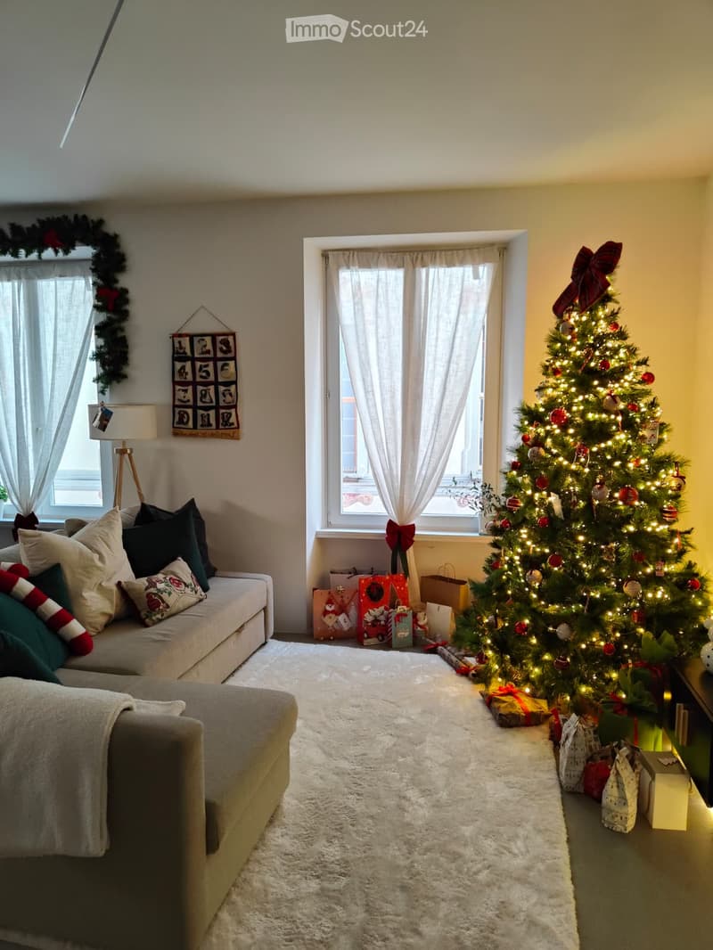 Cozy living room with a large Christmas tree, sofa, chair, and decorative pillows. The room features large windows with curtains and a white shag rug.