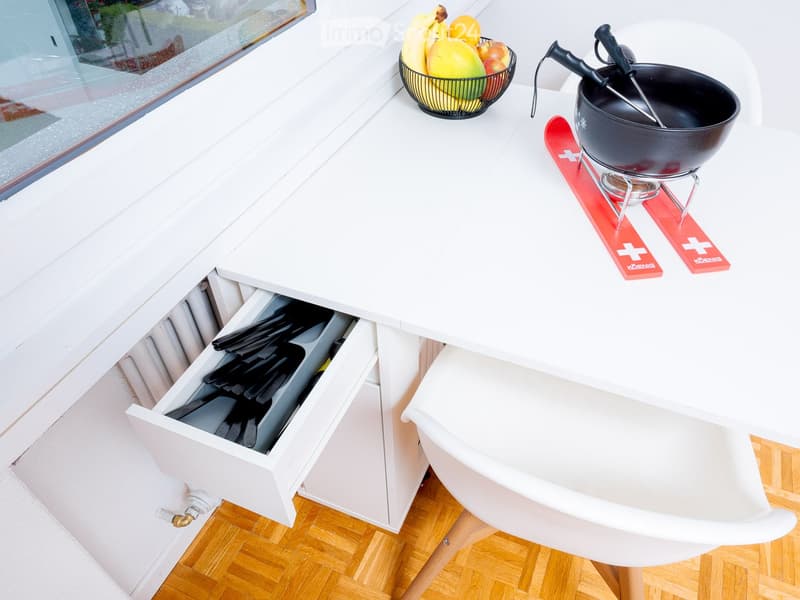 table with a bowl of fruits and kitchen utensils, skis on top, white cabinet under the table, white chairs, wooden floor