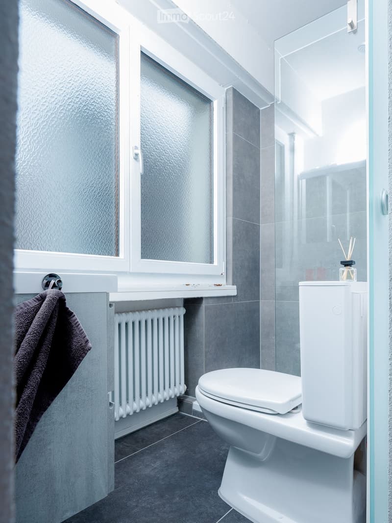Bathroom with white walls, tiled floors, glass shower, white toilet, white radiator, and gray tiles