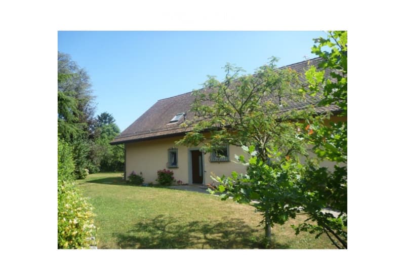 Single-story house with beige walls, brown roof, and small dormer window. The house is surrounded by a well-maintained lawn and trees.