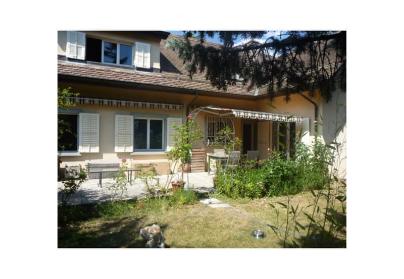 A well-maintained garden area in front of a two-story house with a tiled roof and white shutters. The garden features a small patio with outdoor furniture, potted plants, and a small dog on the grass.