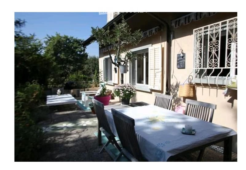 Outdoor patio area with a dining table, chairs, potted plants, and a view of a house with shutters and decorative railings.