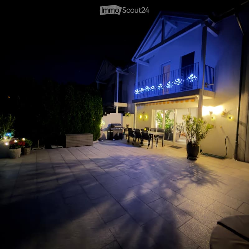 Well-lit outdoor patio with a dining set, grill, potted plants, and string lights on the balcony of a modern house.
