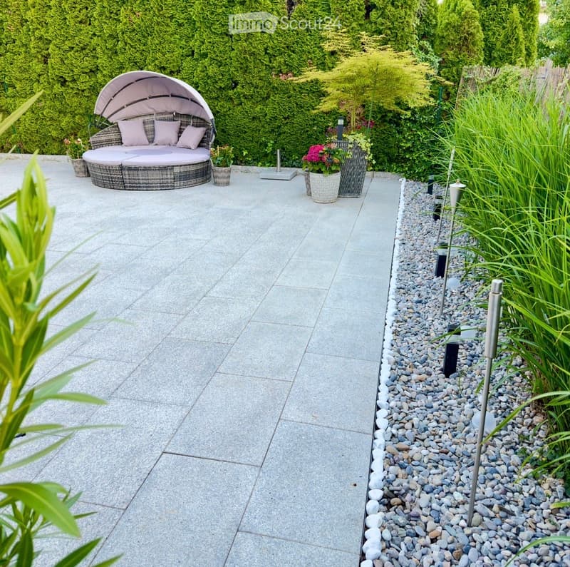 A modern patio with a round wicker daybed, cushions, and surrounding potted plants, bordered by gravel and lush greenery.