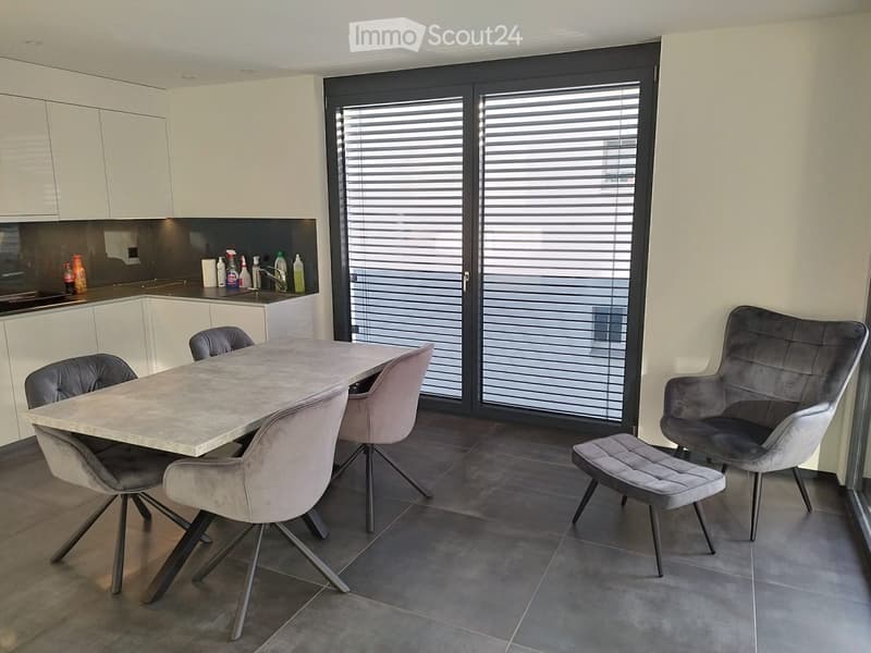 Modern kitchen and dining area with white cabinets, a marble dining table, four chairs, a single armchair, and a small stool. The space features dark tiled flooring and large windows with blinds.