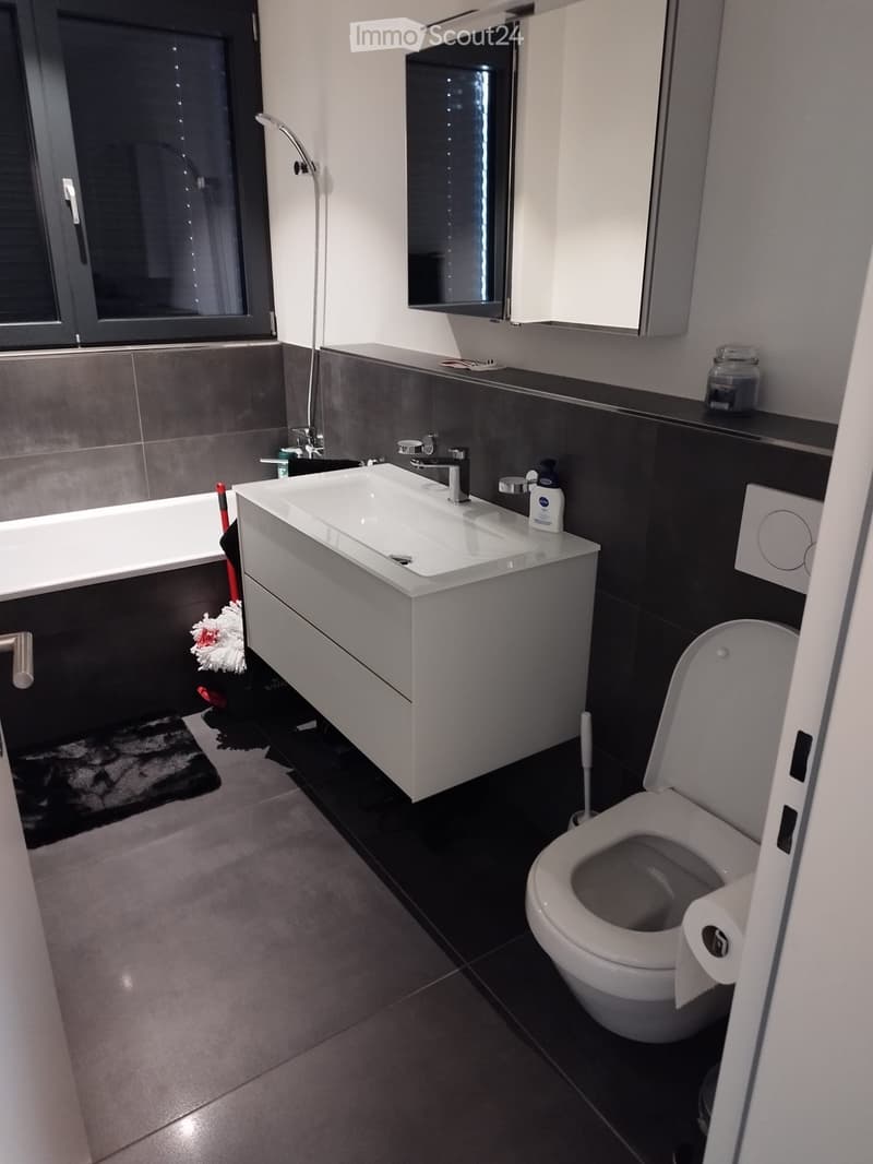 Modern bathroom with a white sink, chrome faucet, toilet, bathtub, and a large mirror above the sink. Dark tiles line the lower half of the walls, and a small jar and bottle sit on the sink counter.