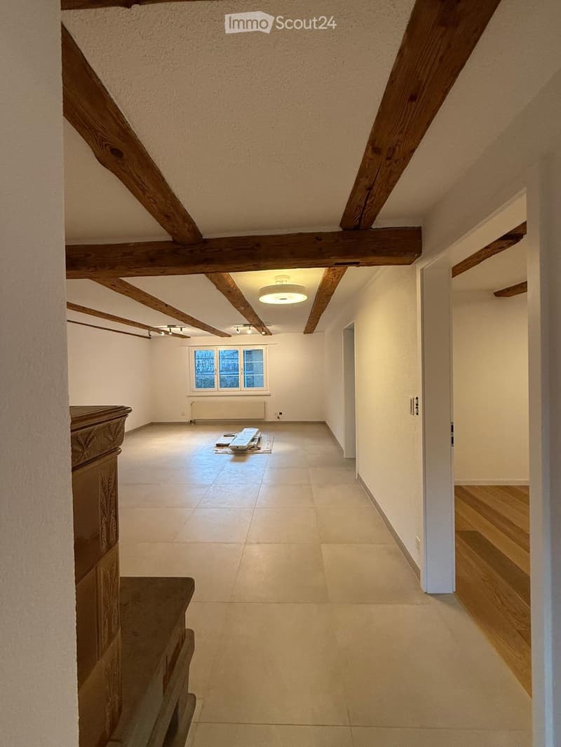 Empty room with exposed wooden beams, tiled floor, and a window with a radiator below it. The room has a simple, clean design with white walls and a recessed ceiling light.
