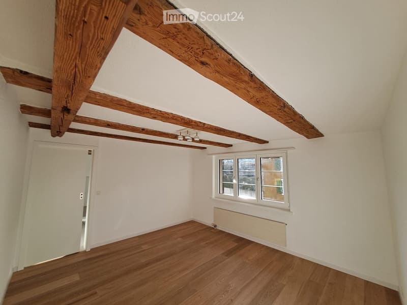 Empty attic room with wooden beams, wooden flooring, white walls, and a window with multiple panes.