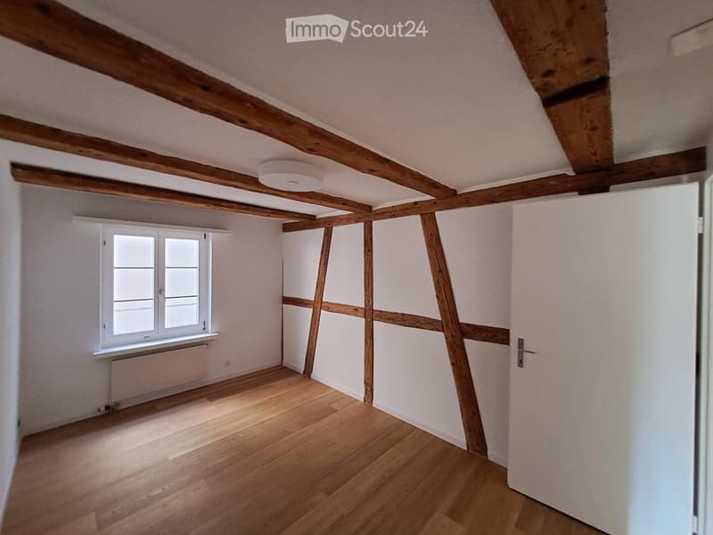 Empty attic room with wooden beams, wooden floor, white walls, and a window with white frame. No furniture or equipment present.