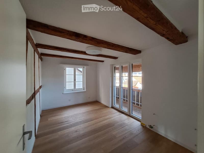 Empty room with wooden floor, exposed wooden beams on the ceiling, white walls, and a large window with white frames. A sliding glass door leads to a balcony or terrace area.
