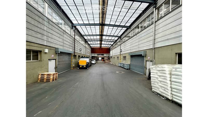 Large industrial warehouse with a high, translucent roof, concrete floors, and multiple loading docks. Visible elements include pallets, a small vehicle, and stacked materials.