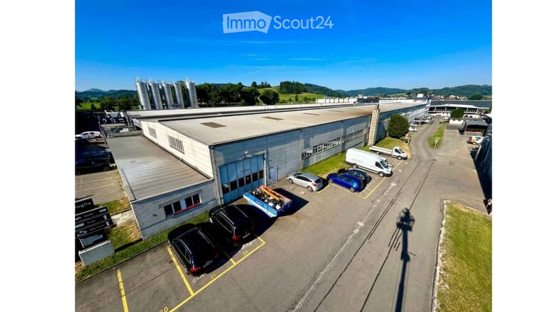 Aerial view of a large industrial building with a flat roof, surrounded by a parking lot with several cars and vans. The building has a modern design with large windows and a brick facade. There are additional structures and storage tanks visible in the b