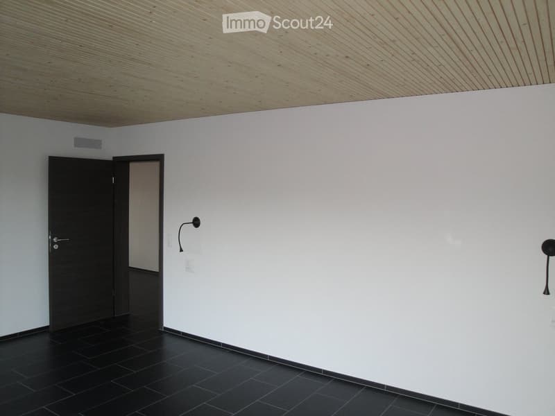 Empty room with white walls, black tiled floor, and a wooden slatted ceiling. Features a black door, wall-mounted light fixture, and a small vent on the wall.