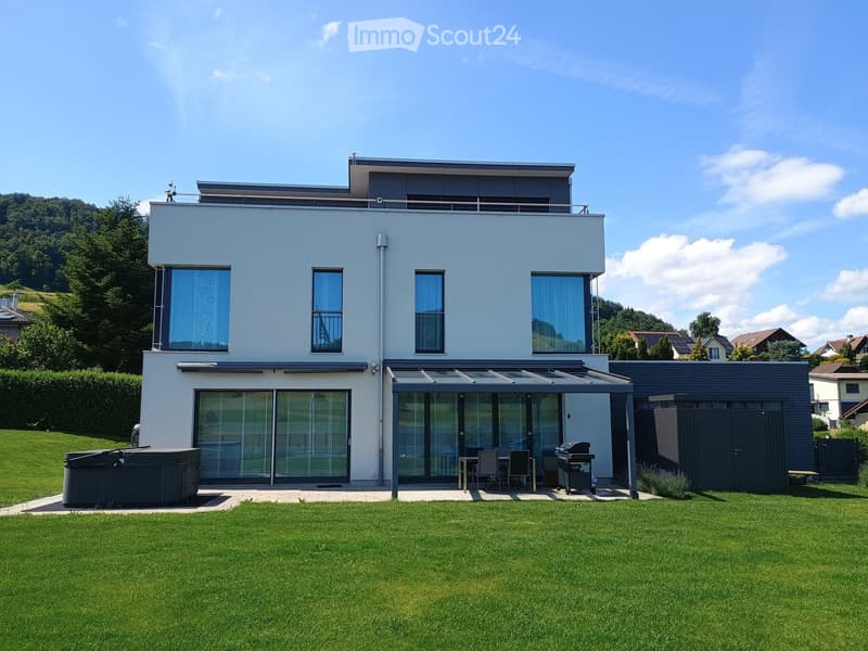 Modern two-story house with light blue exterior, large windows, covered patio area, outdoor seating, grill, and hot tub in a well-maintained lawn.