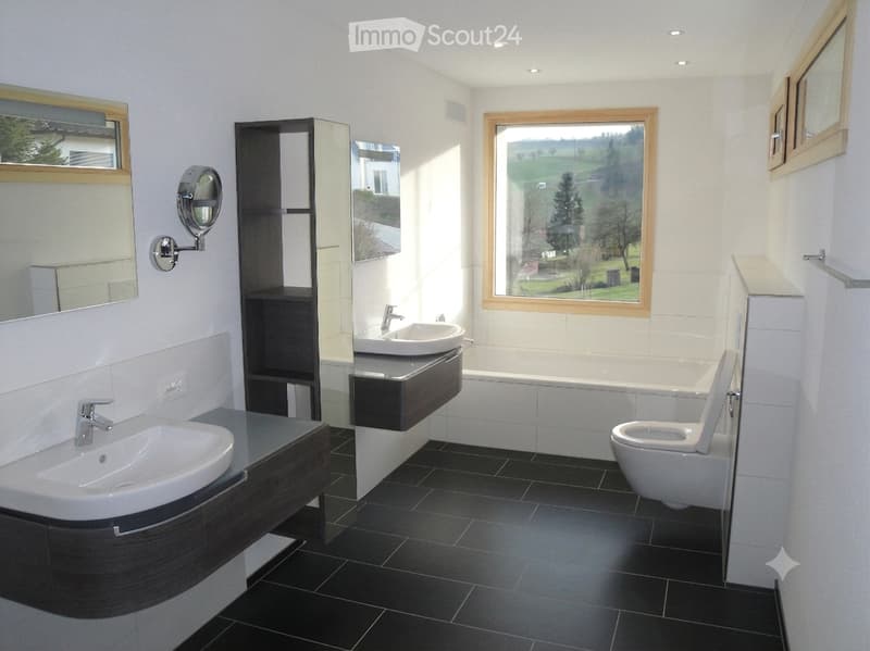 Modern bathroom with black tiled floor, white walls, two sinks, a toilet, a bathtub, and a large window offering a scenic view.