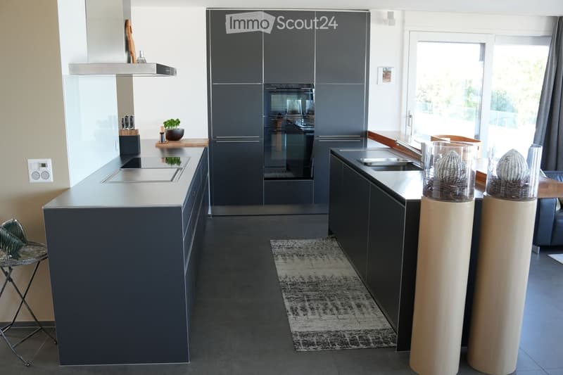 Modern kitchen with dark grey cabinets, stainless steel appliances, island with integrated stove, sink, and seating area. Features a small potted plant, knife block, and decorative columns.