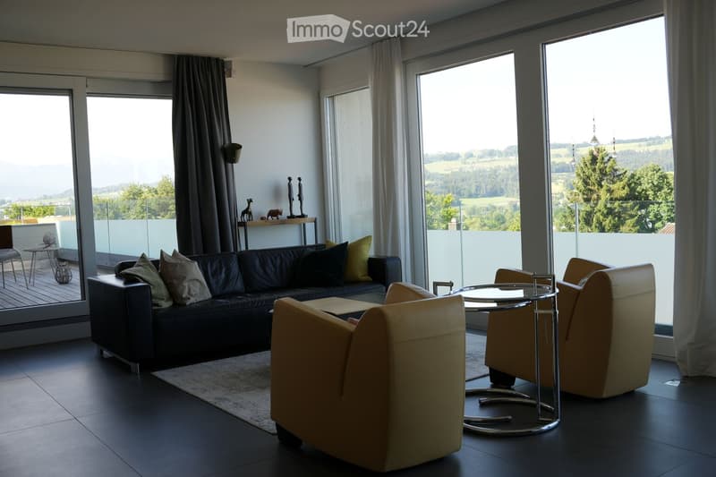 Modern living room with large windows offering a scenic view, featuring a black leather sofa, cream armchairs, a round glass coffee table, and a small side table with decorative items.