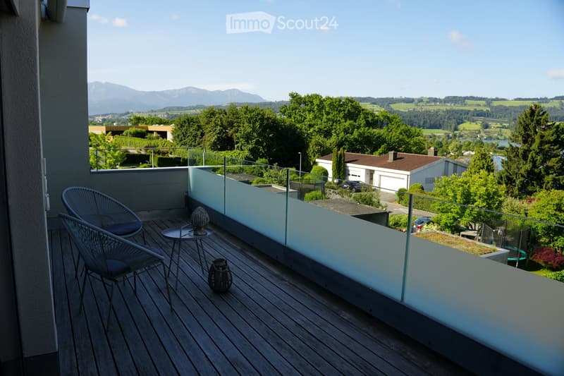 Balcony with wooden decking, glass railing, two chairs, small table, and decorative vases, overlooking a scenic landscape with mountains and greenery.