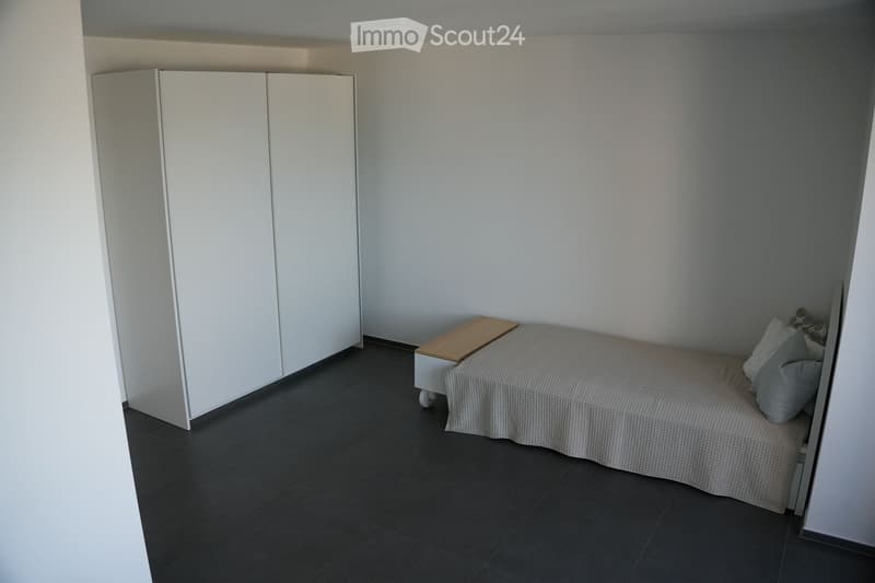 Modern, minimalist bedroom with a single bed, white wardrobe, and a small nightstand. The room features dark tiled flooring and light-colored walls.