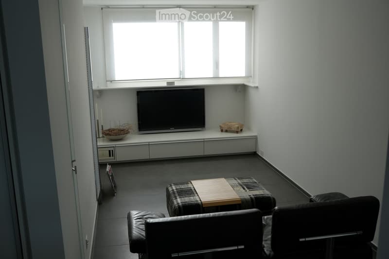 Modern living room with dark tiled flooring, white walls, and large windows. Features a sofa, coffee table, television on a white cabinet, and decorative items including a bowl and candles.