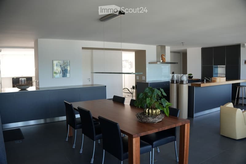 Modern open-plan kitchen and dining area with dark blue cabinetry, a wooden dining table with black chairs, a kitchen island, and a small seating area with a cream-colored armchair.