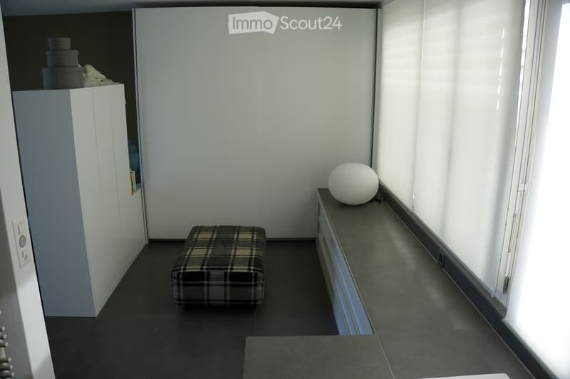 A small, modern hallway with a dark tiled floor, white walls, a plaid-patterned stool, a white spherical decorative object on a cabinet, and a shelf with books and boxes.