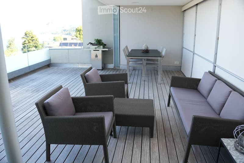 Balcony with wooden decking, wicker furniture including a sofa, chairs, ottoman, and a small table with chairs.