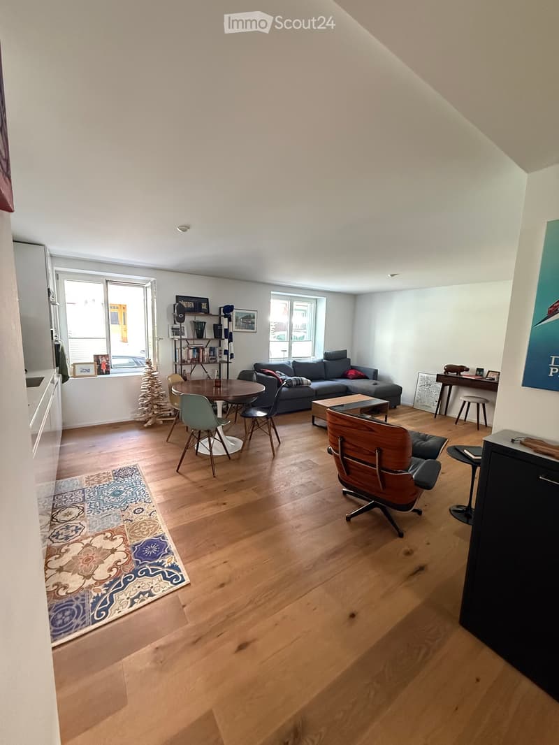 Bright living room with wooden flooring, large windows, a grey sectional sofa, a round dining table with chairs, a wooden armchair, a small shelf with books and decor, and a patterned rug.