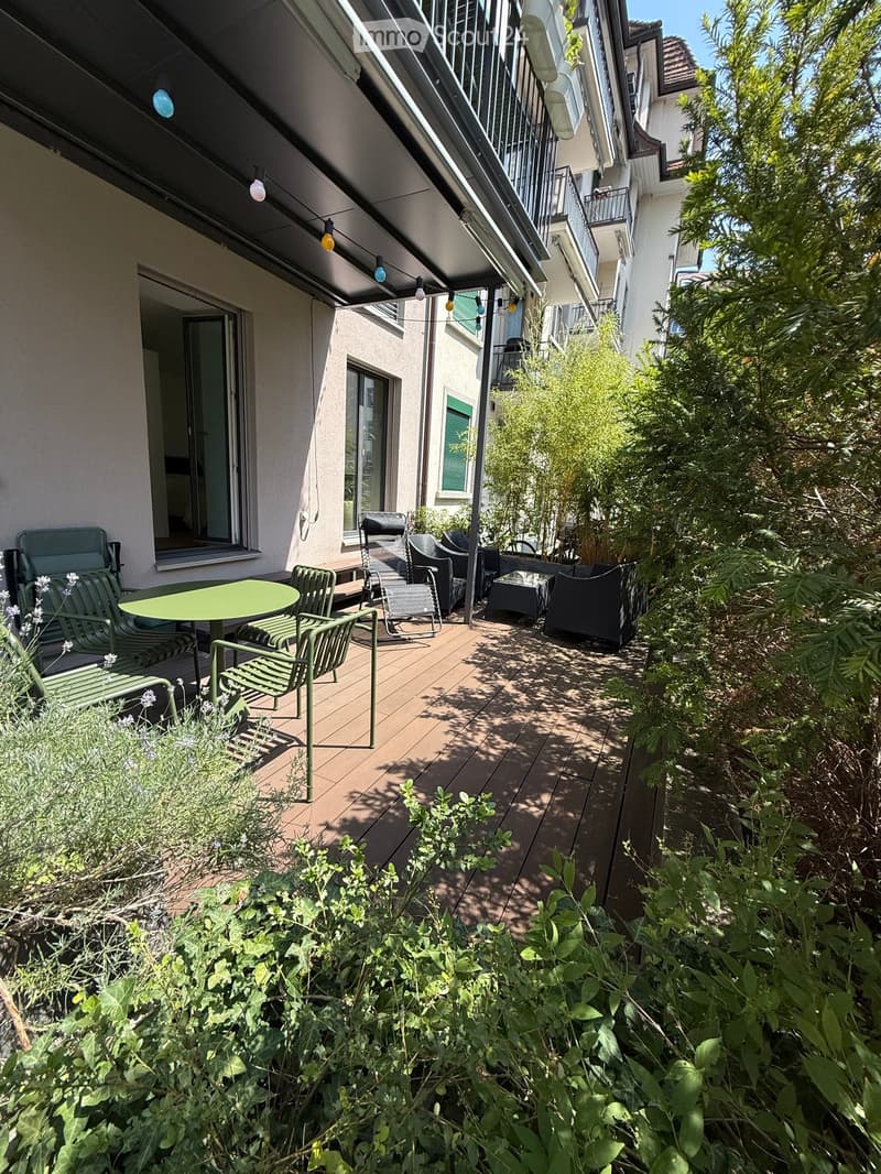 Balcony with wooden decking, green table, and several chairs, surrounded by lush greenery and plants.