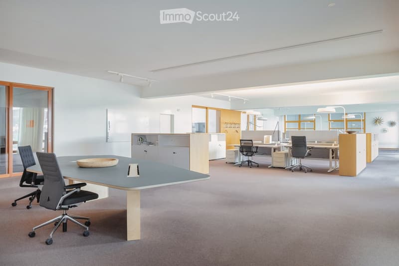 Modern office space with a large central table, chairs, desks, cabinets, and a screen on the wall.