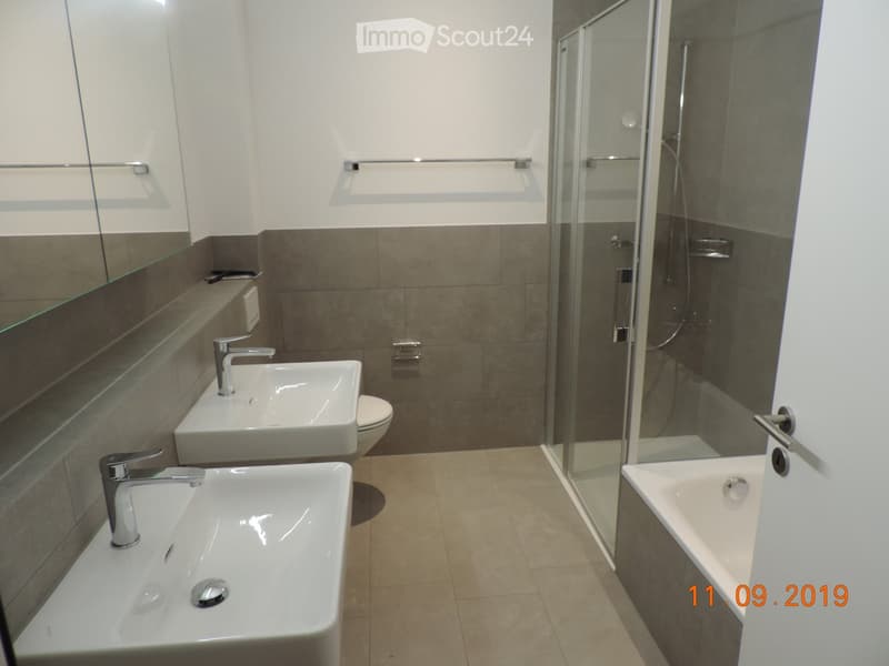 Modern bathroom with two sinks, a toilet, a bathtub, and a glass-enclosed shower. Features include a large mirror, modern faucets, and tiled walls.