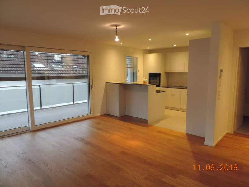 Open-plan living and dining area with wooden flooring, white kitchen cabinets, and a large sliding glass door leading to a balcony.