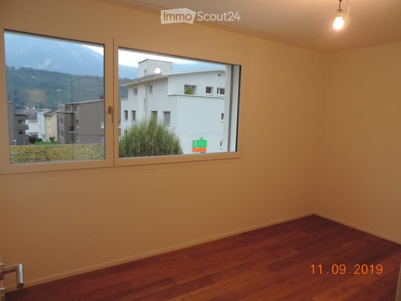 Empty room with wooden flooring and large windows offering a view of a building and mountains.
