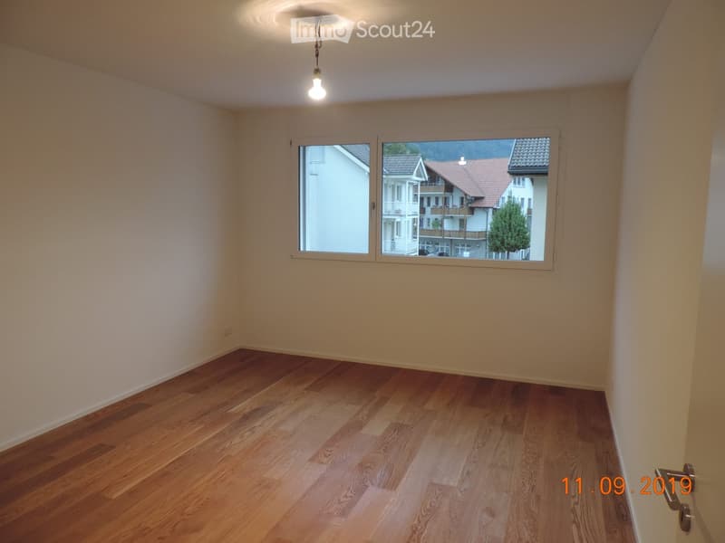 Empty room with wooden flooring, white walls, and large windows offering a view of neighboring buildings.