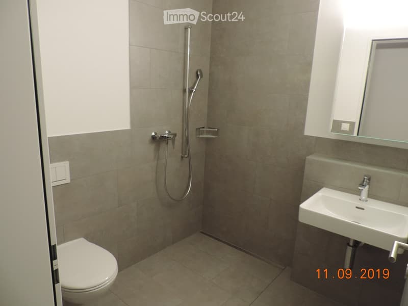 Modern bathroom with beige tiled walls and floor, featuring a toilet, a sink with a mirror, and a shower with a handheld sprayer.