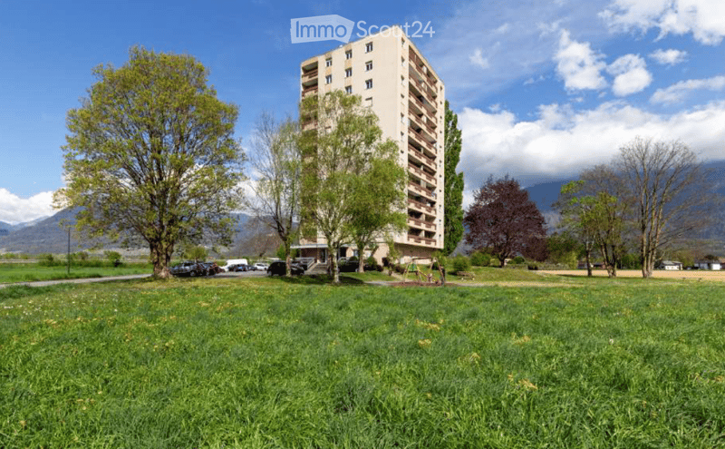 A multi-story residential building with balconies, surrounded by a green lawn, trees, and a playground area.