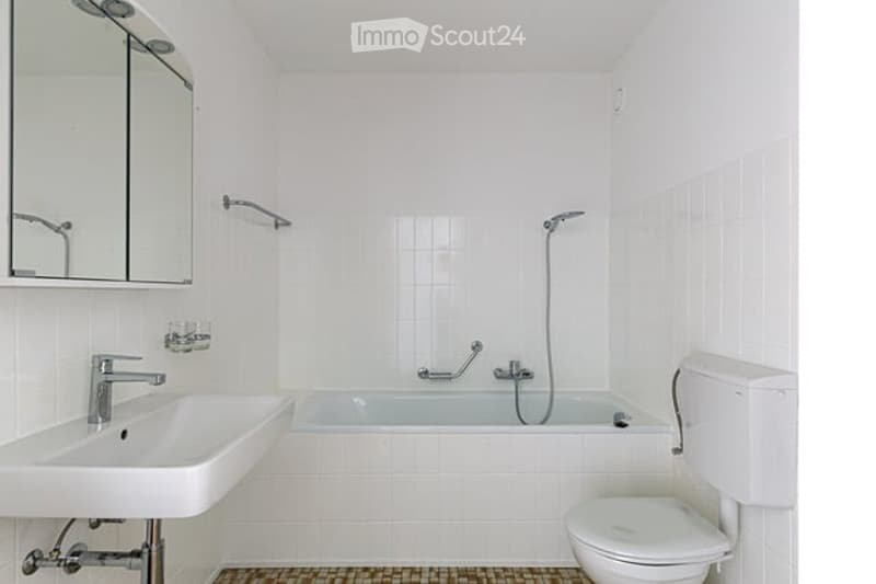 Modern, white-tiled bathroom with a sink, bathtub, toilet, and mirror. The space is clean and minimalistic with a showerhead visible above the bathtub.