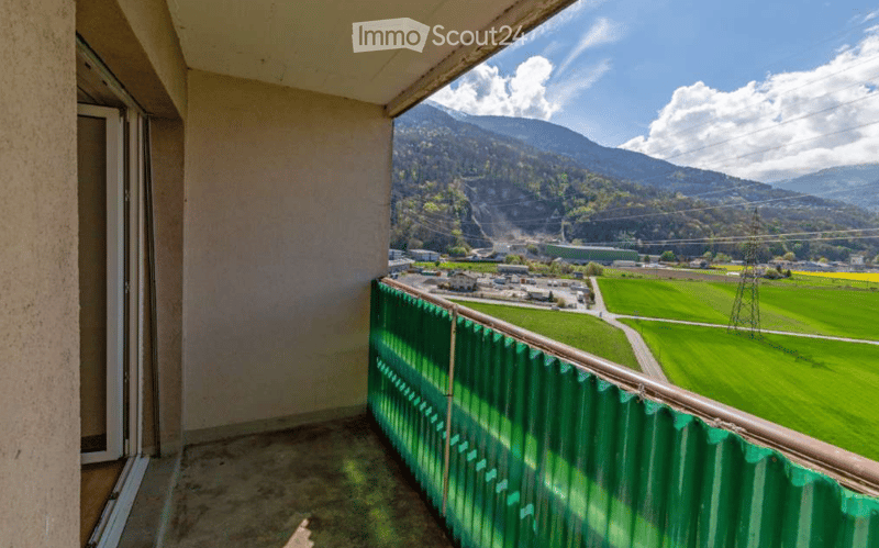 Balcony with green railing, overlooking a scenic valley with mountains, fields, and scattered buildings.