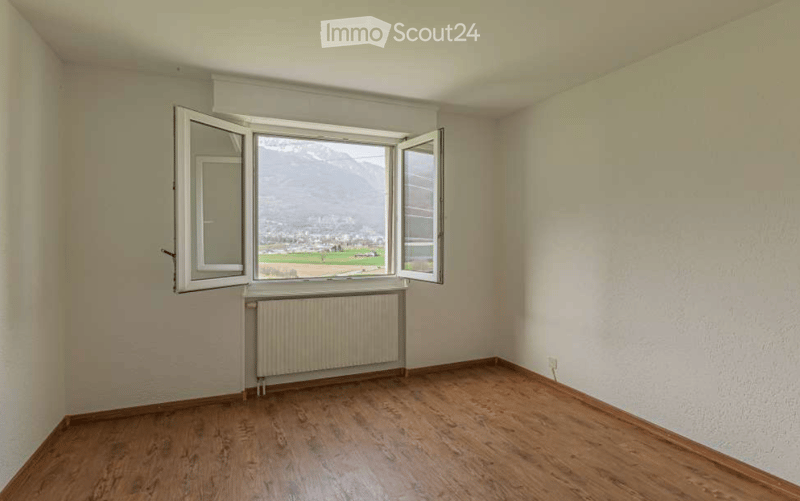 Empty room with wooden flooring, white walls, large window with open shutters, radiator, and a scenic mountain view.