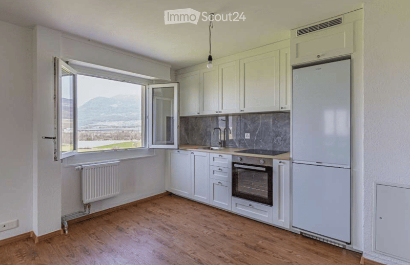 Modern kitchen with white cabinets, wooden flooring, and large windows offering a scenic view. Features include a refrigerator, oven, sink, and faucet.