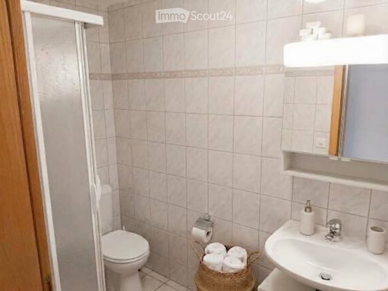 A small bathroom with beige tiled walls and floor, featuring a toilet, sink, mirror, and a woven basket with towels.