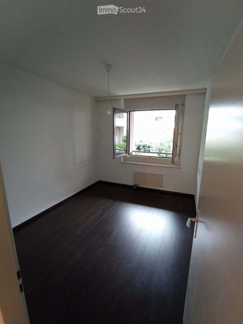 Empty room with wooden flooring, white walls, and a large window with open shutters allowing natural light to enter.