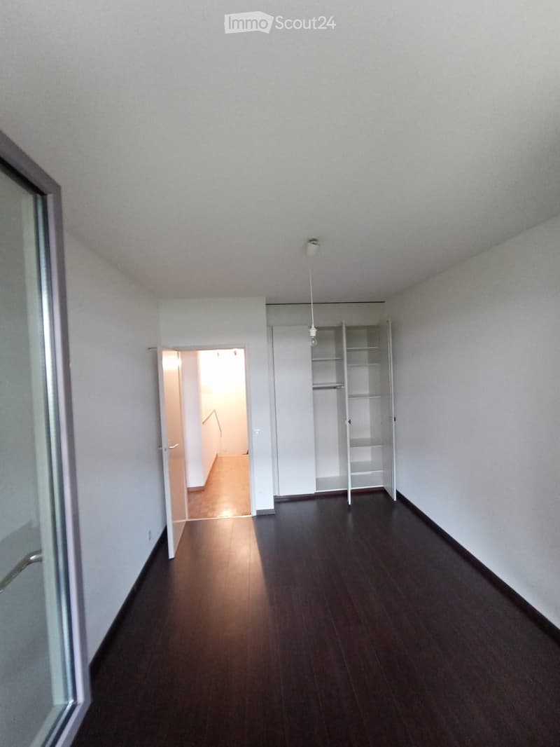 Empty hallway with dark wooden flooring, white walls, and a single open closet with empty shelves. A doorway leads to another room with light-colored flooring.
