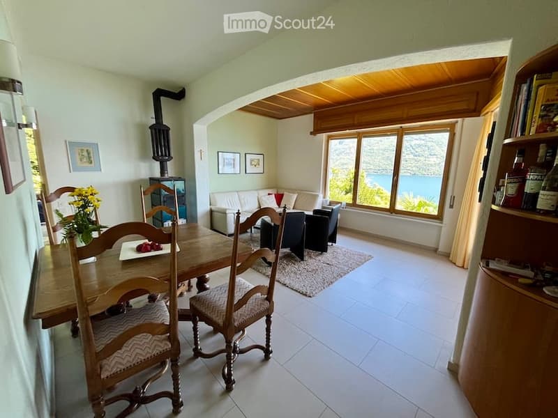 Dining area with table, chairs, rug, side table with flowers, white walls, white tile floor, fireplace, shelf with books and bottles, chairs, couch, large windows with scenic view, lamps