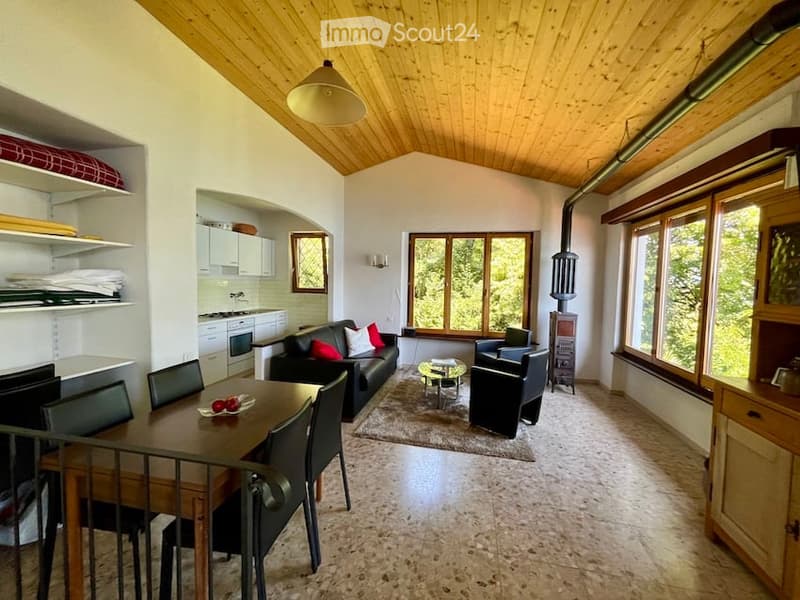 wooden ceiling, white walls, a large window, a black couch with pillows, a dining table with four chairs, a rug, a cabinet, a shelf, a stove and cabinets, a lamp, a glass table, a black chair
