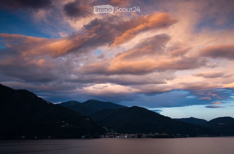 Panoramic landscape with mountains, a town, and a serene lake under a dramatic sky