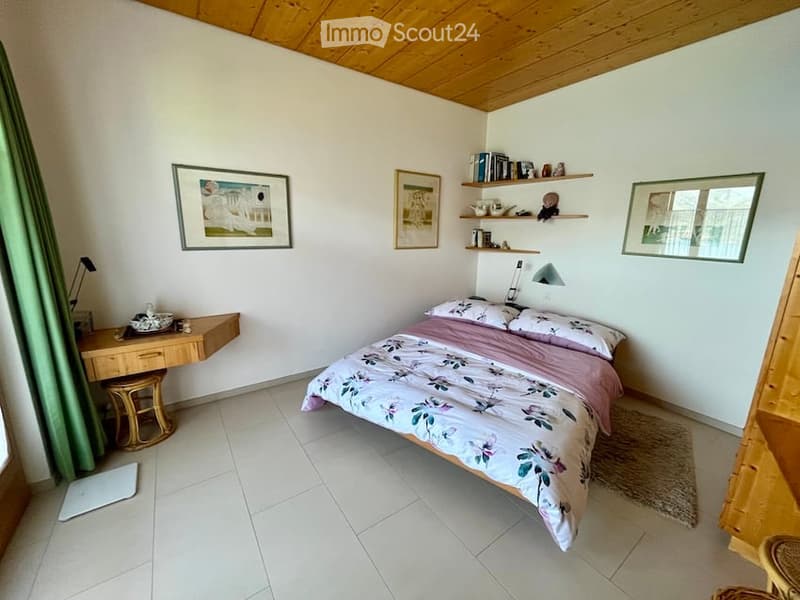A cozy bedroom with a bed, a desk, a rug, and picture frames on the wall. Wooden ceiling and floor.